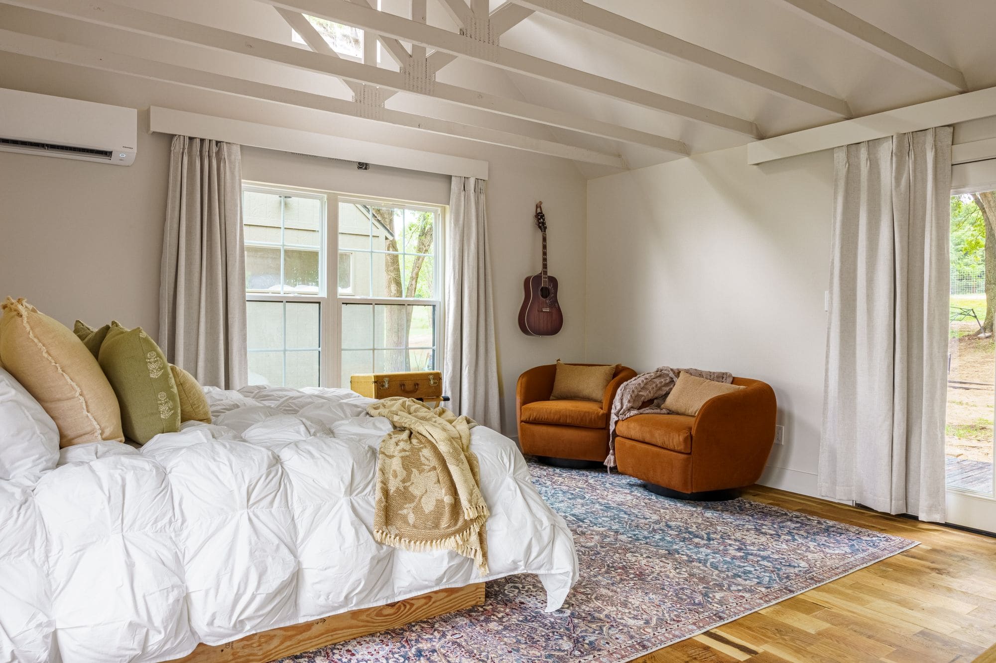 Cabin bedroom with a bed, white duvet, a rust leather chair and a guitar on the wall under a beamed ceiling