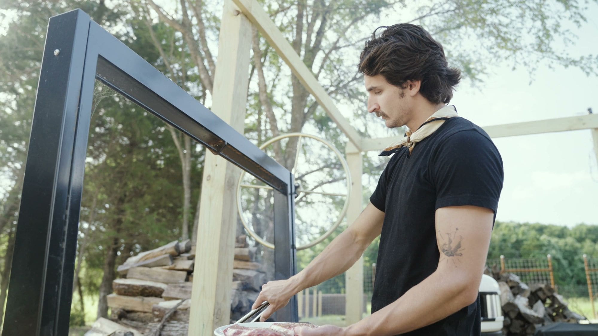 Tending the wood-fired grill with the trees behind